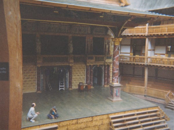 A photo showing the stage of Shakespeare's Globe Theatre. Two mean sit on the empty stage, appearing deep in discussion